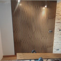 Bizland - Montaż dekoracyjnych paneli ściennych z drewna. Częściowo wykończona ściana z drewnianymi lamelami, drabina i narzędzia na podłodze w trakcie prac wykończeniowych.