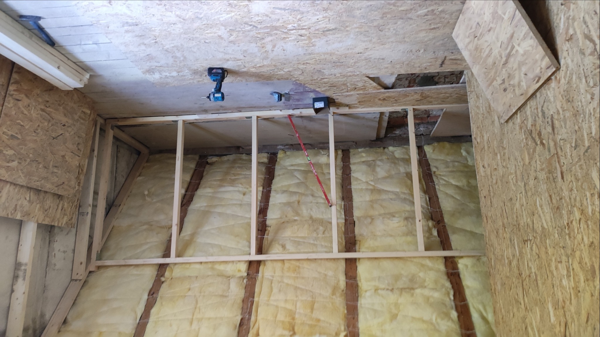 Skośna ściana z izolacją z wełny mineralnej, drewnianą konstrukcją szkieletową i częściowo zamontowaną płytą OSB. W tle widoczne narzędzia: poziomica i wkrętarka.