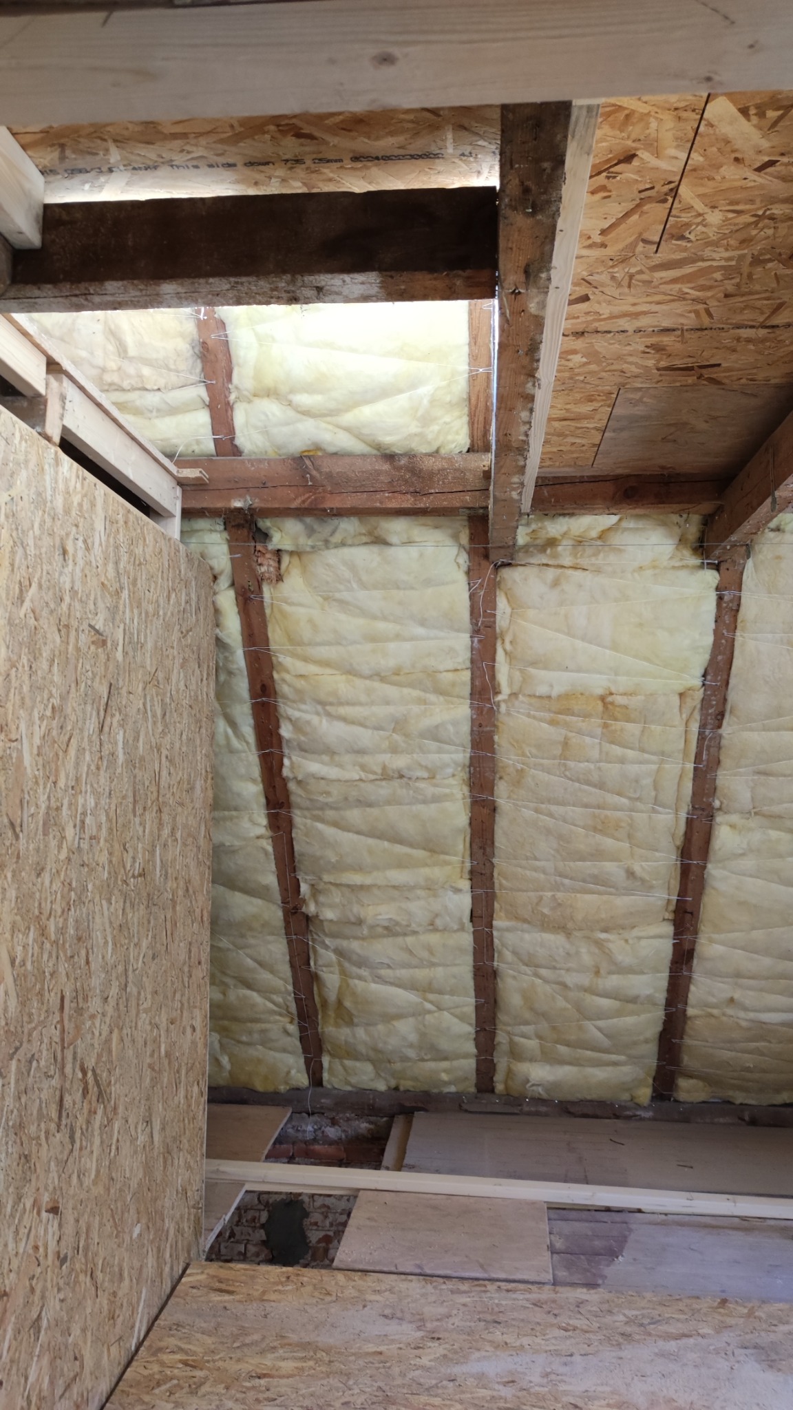 Izolacja poddasza wełną mineralną przymocowaną drutem do drewnianych belek, widoczne płyty OSB na podłodze i ścianie. Prace wykończeniowe.