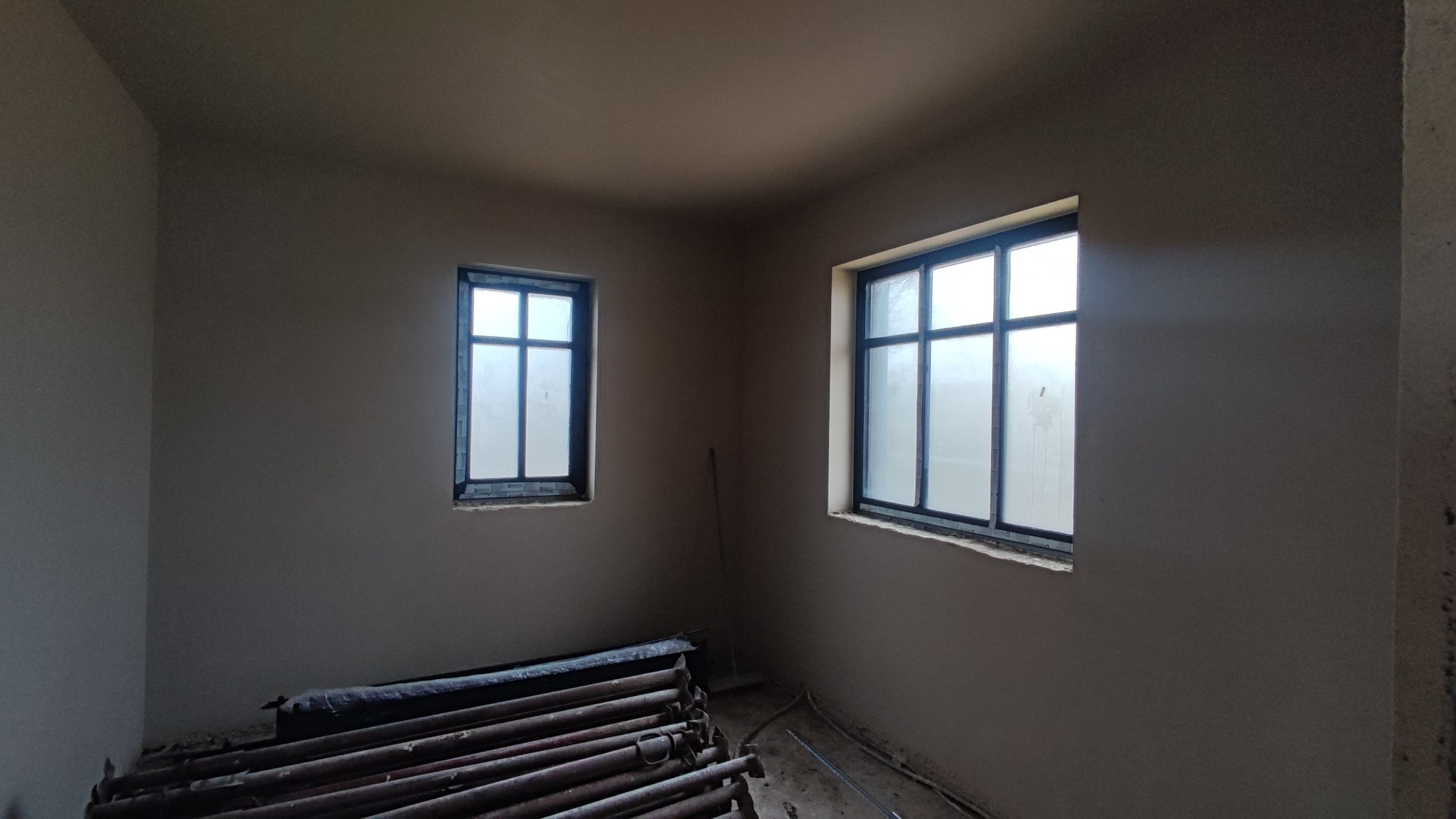 Pokój po tynkowaniu maszynowym: dwie okna z czarnymi ramami, szare ściany, stos rusztowań leżący na podłodze. Widok na świeżo otynkowane ściany.