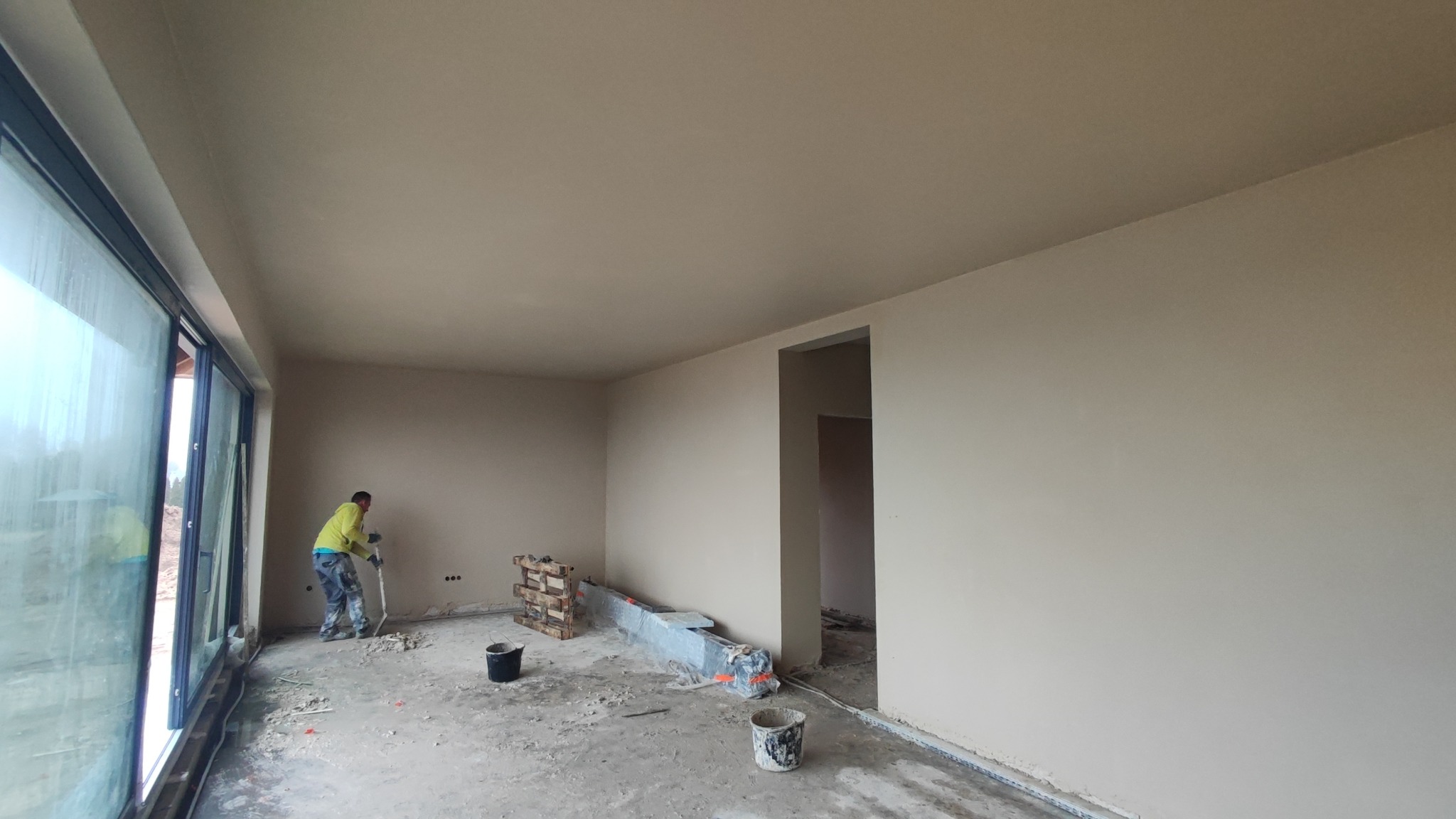 Wnętrze po tynkowaniu maszynowym: gładkie ściany w beżowym odcieniu, duże okno z widokiem na zewnątrz, pracownik z narzędziem na podłodze. Widoczne ślady prac.