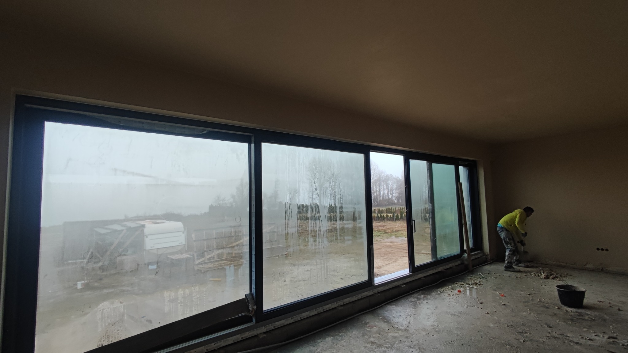 Wnętrze w trakcie prac wykończeniowych: pracownik szpachluje ścianę. Duże okno z widokiem na zewnątrz, na podłodze resztki materiałów budowlanych, widoczne ślady szpachlowania.