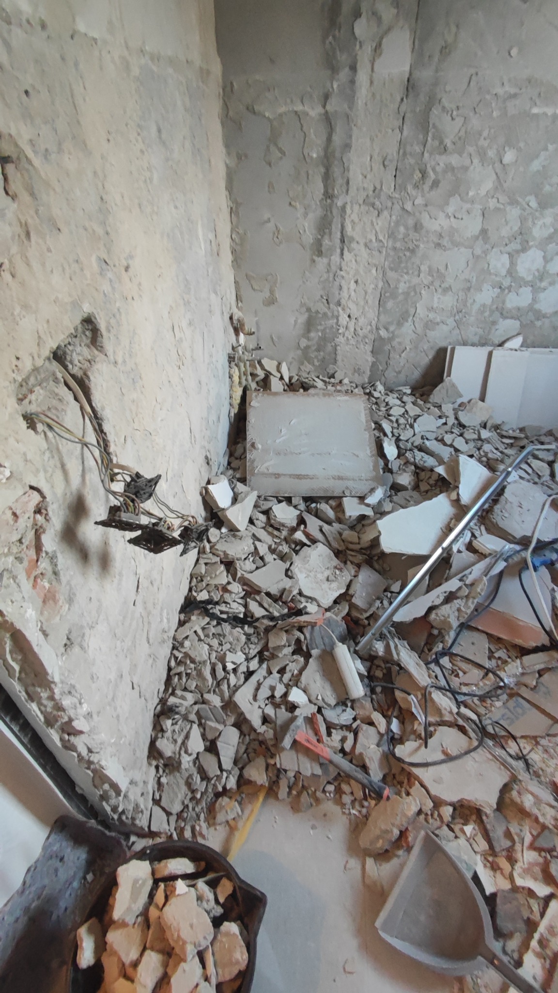 Wnętrze w trakcie remontu: odsłonięte przewody elektryczne, gruz i narzędzia na podłodze. Widoczne ślady po skuciu tynku na ścianach.