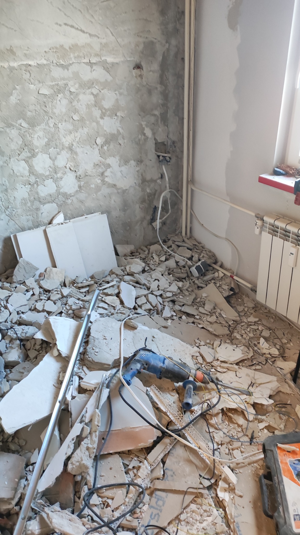 Demontaż ściany: gruz, płyty, wiertarka udarowa. Ściana w tle z widocznymi instalacjami. Grzejnik pod oknem. Etap przygotowawczy do dalszych prac.