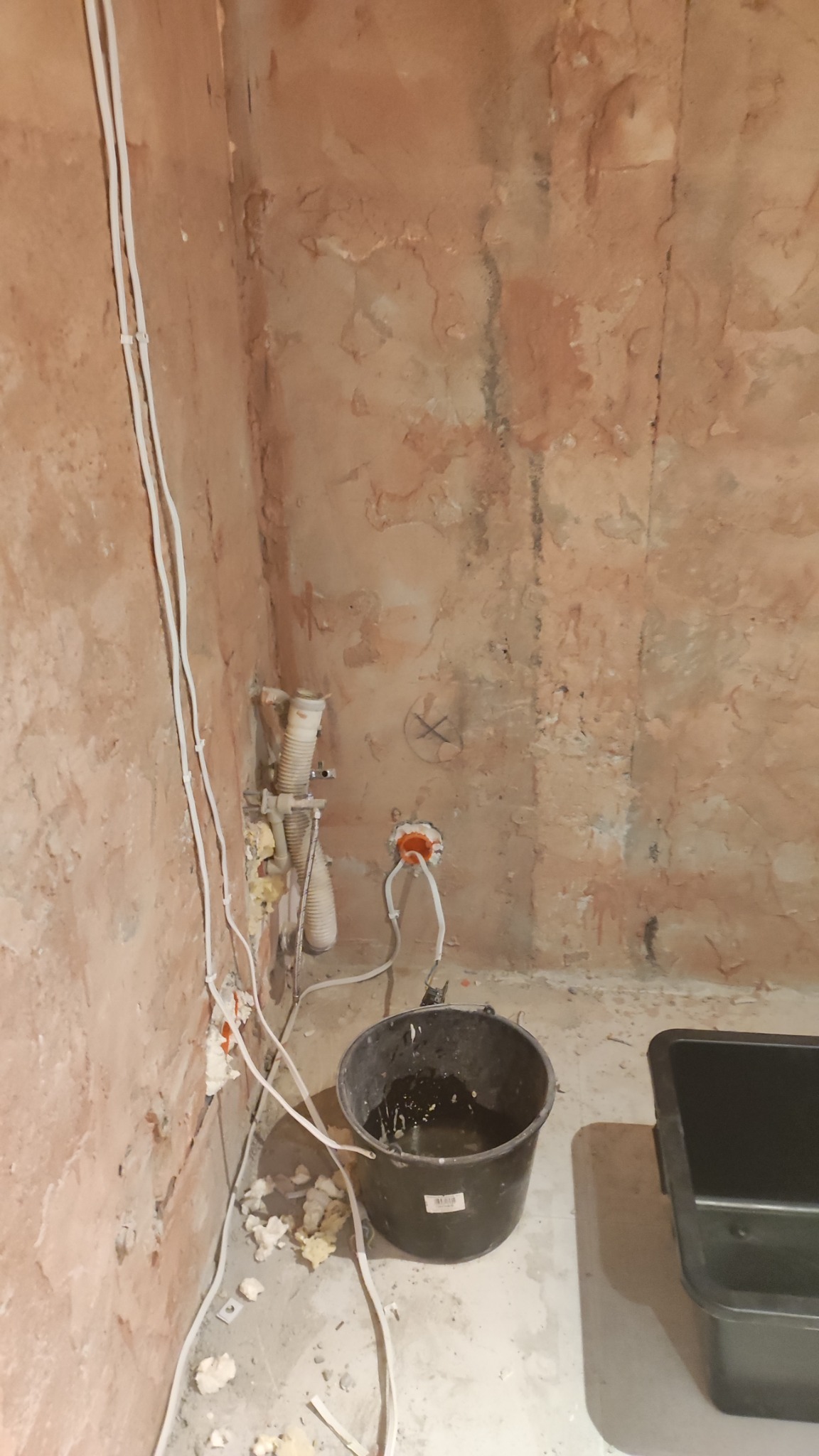 Surowa ściana w trakcie remontu: widoczne przewody elektryczne, rury, wiadro i pojemnik. Ściana o nierównej fakturze, przygotowana do dalszych prac wykończeniowych.