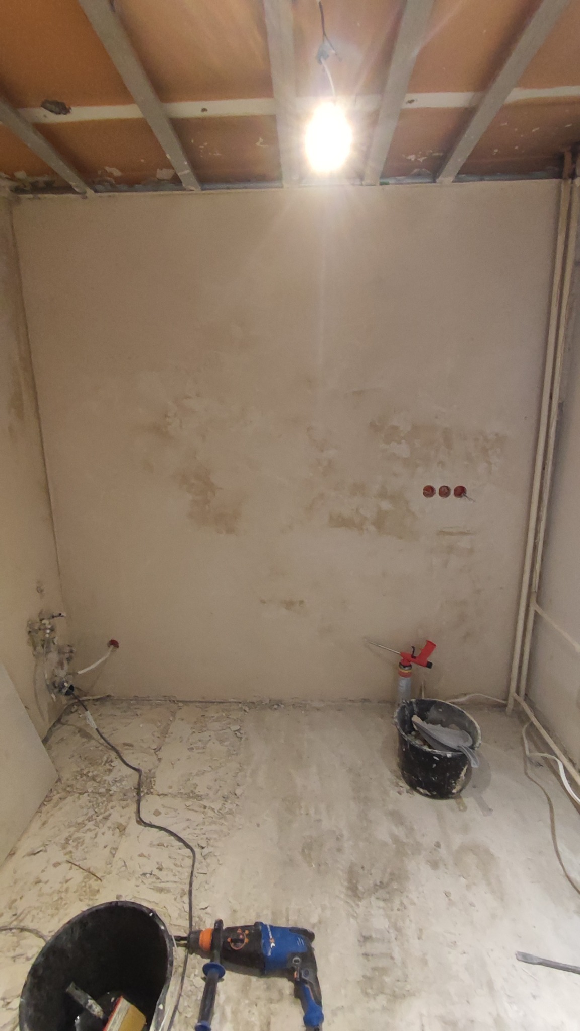 Wnętrze w trakcie prac przygotowawczych: ściany po nałożeniu warstwy tynku, widoczne narzędzia (wiertarka, wiadro) i instalacje. Sufit z odsłoniętą konstrukcją.