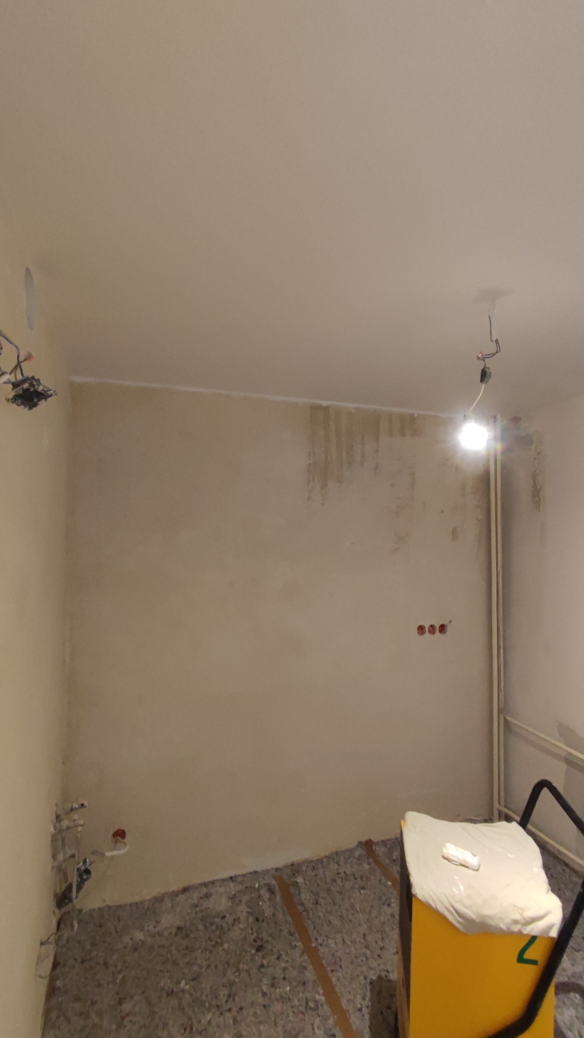 Pokój w trakcie prac wykończeniowych: ściany po szpachlowaniu, widoczne instalacje elektryczne i hydrauliczne, osłonięty sprzęt budowlany na podłodze.