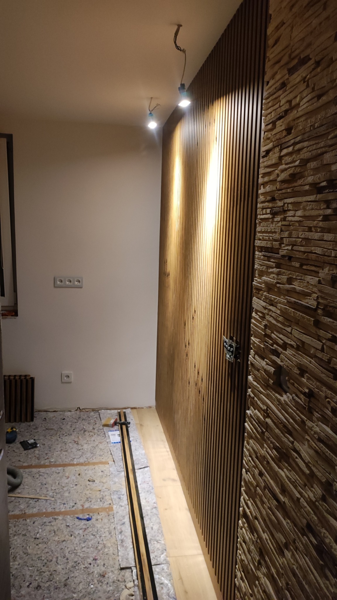 Wnętrze w trakcie prac wykończeniowych: panele ścienne drewniane i kamienne na ścianie, podłoga przygotowana do montażu, widoczne gniazdka elektryczne.