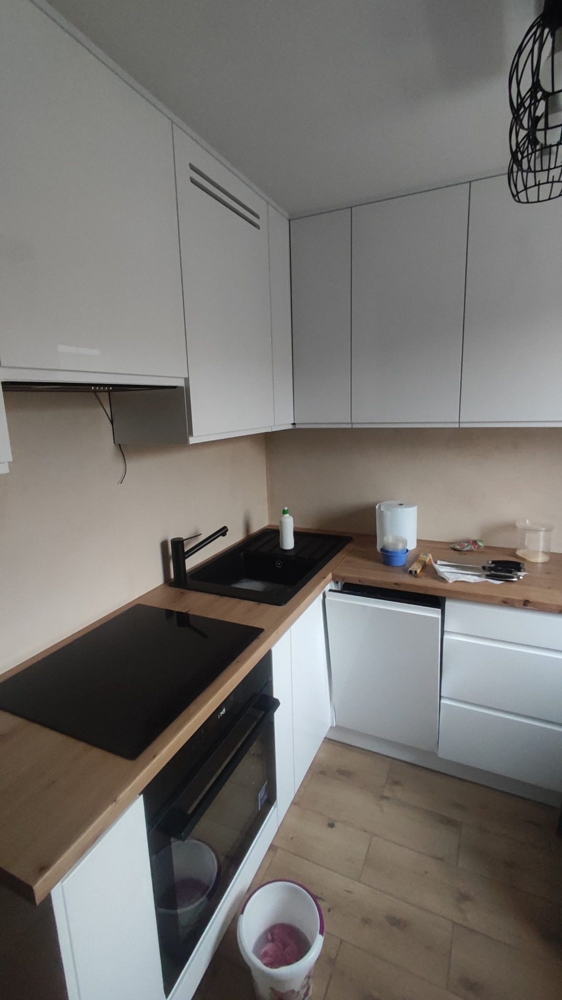 Nowoczesna kuchnia w bieli i drewnie: zabudowa meblowa z czarnym zlewem i płytą indukcyjną, drewniany blat, minimalistyczna lampa. Widoczny fragment podłogi i wiadro.