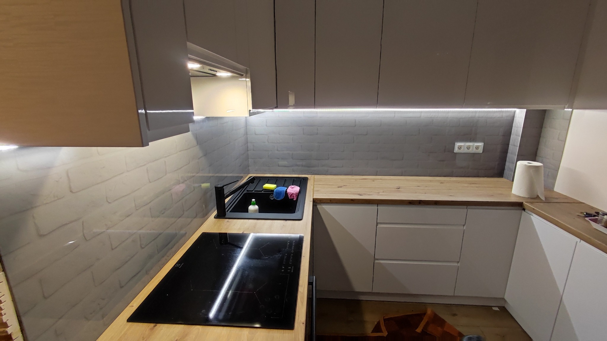 Nowoczesna kuchnia w szaro-białej tonacji z drewnianym blatem, czarnym zlewem i płytą indukcyjną. Ściana wykończona imitacją cegły, oświetlenie LED pod szafkami.