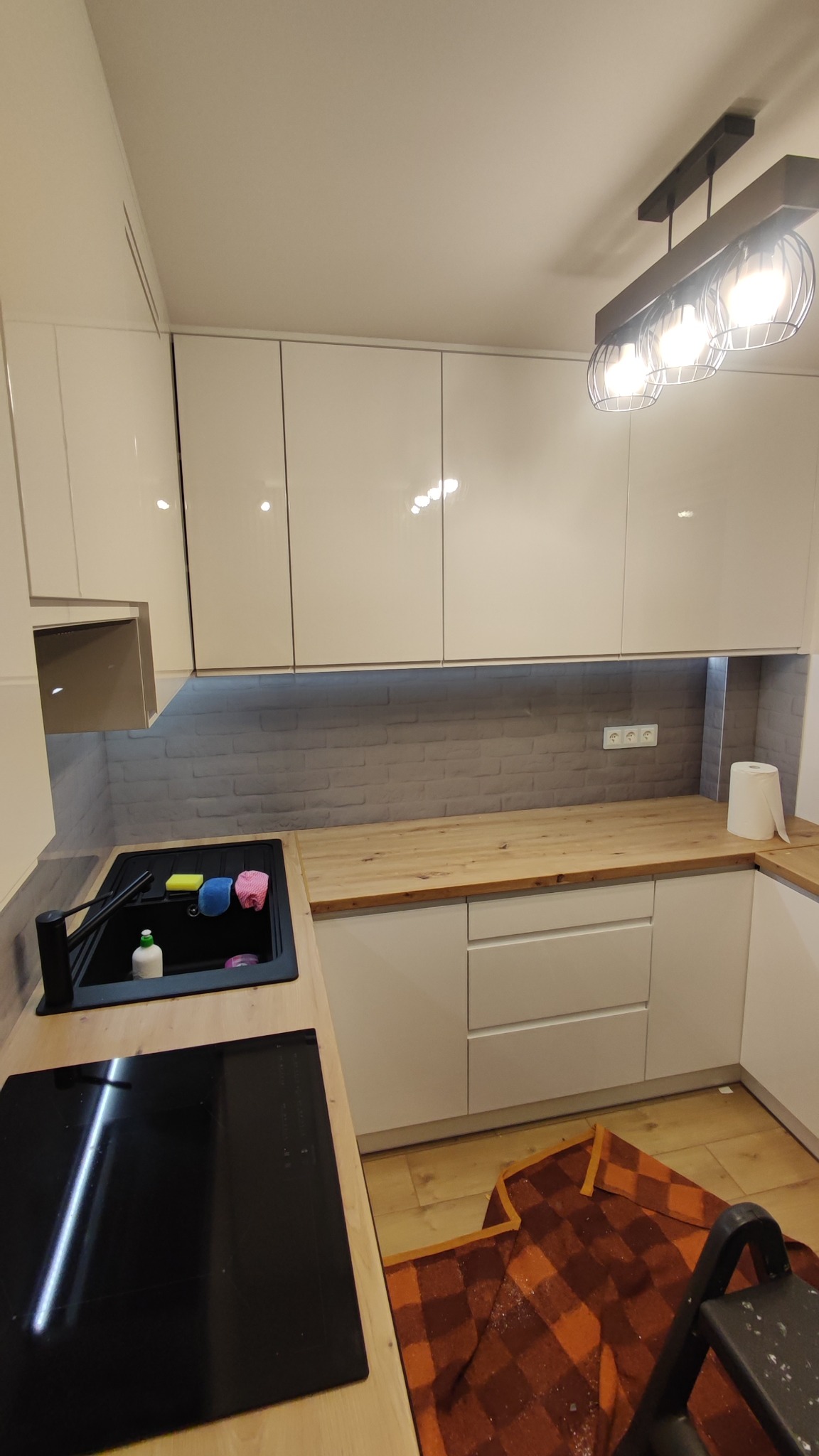 Nowoczesna kuchnia z białymi, błyszczącymi szafkami i drewnianym blatem. Uwagę zwraca szara ściana imitująca cegłę oraz czarny zlew z akcesoriami.