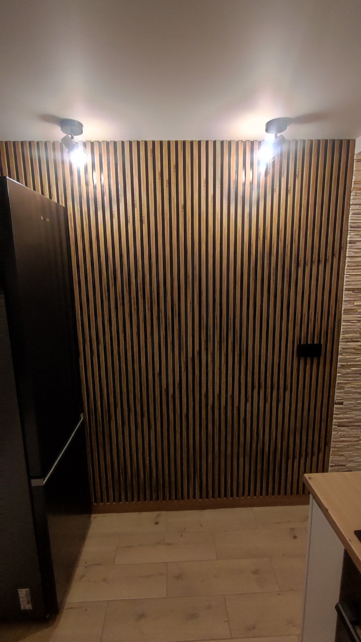 Ściana z drewnianych lameli w kuchni, z widocznymi dwoma lampami sufitowymi i fragmentem lodówki po lewej stronie. Dekoracja wnętrza w nowoczesnym stylu.