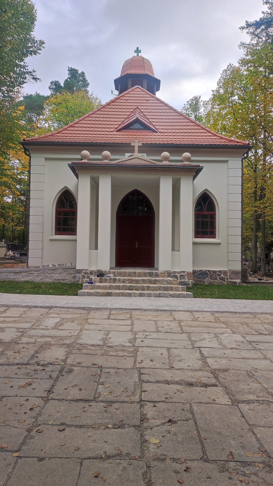 Odnowiona, kremowa kaplica z czerwoną dachówką, widoczna z brukowanego placu, otoczona drzewami z jesiennymi liśćmi. Uwagę przykuwają łukowe okna i drzwi.