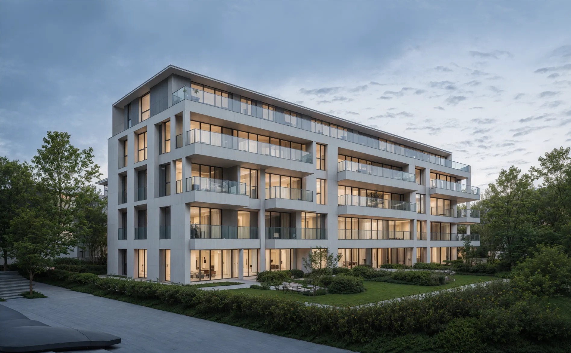 Elewacja nowoczesnego, wielopiętrowego apartamentowca z balkonami i dużymi oknami, otoczonego zielenią. Widoczny fragment chodnika i drzewa. Oświetlenie wieczorne.