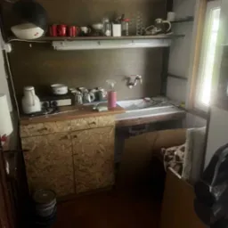 Zaniedbana kuchnia z drewnianymi szafkami, na których stoją naczynia. Nad szafkami półka z kubkami i innymi akcesoriami kuchennymi. W rogu widoczny odkurzacz.