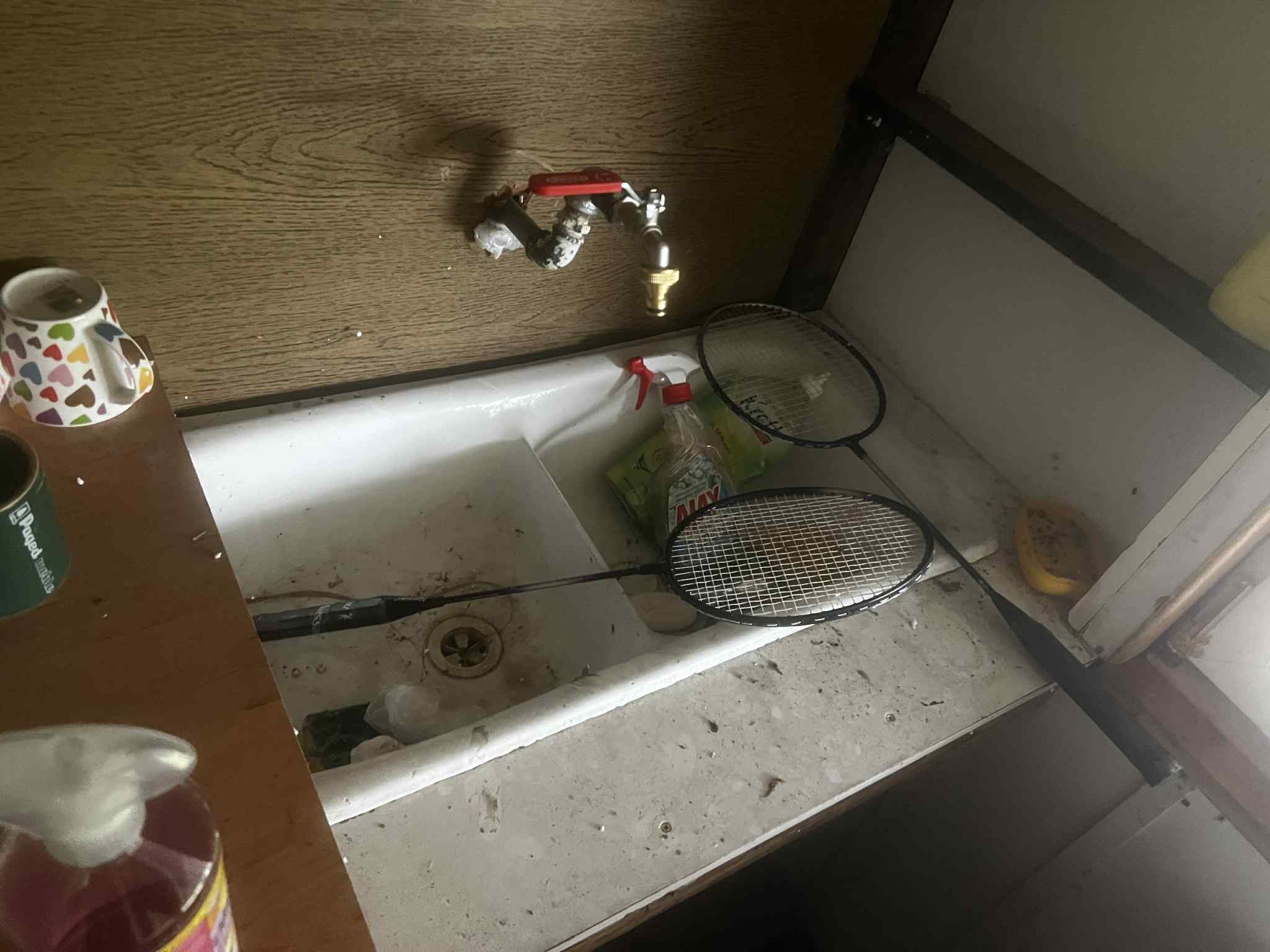 Zaniedbana kuchnia: brudny zlew z kranem, płyn do naczyń, kubek, puszka i rakietki do badmintona. Widoczny bałagan i brak czystości.