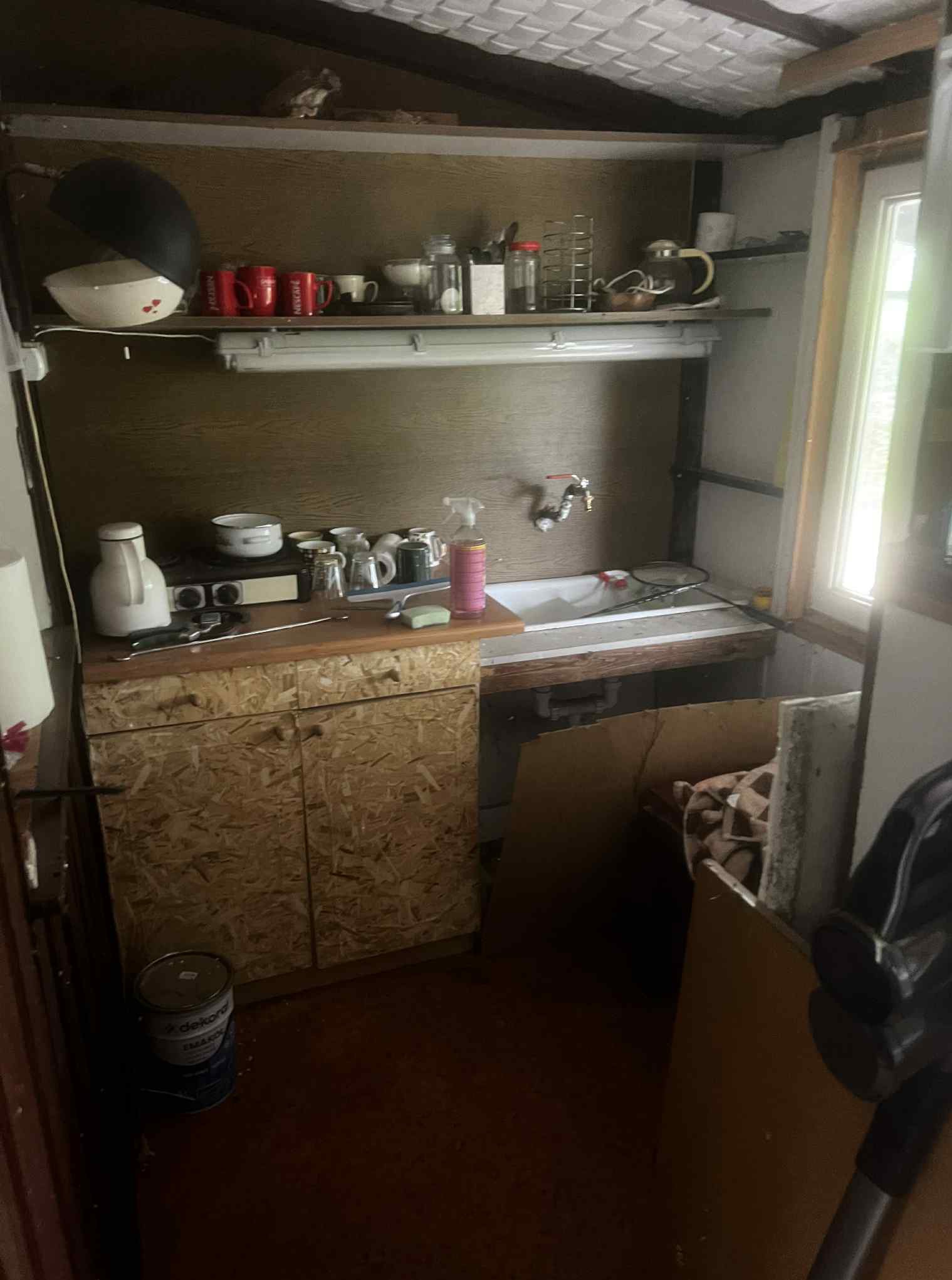 Zaniedbana kuchnia z drewnianymi szafkami, na których stoją naczynia. Nad szafkami półka z kubkami i innymi akcesoriami kuchennymi. W rogu widoczny odkurzacz.