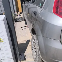 Szary samochód osobowy z otwartą maską, uniesiony na podnośniku, narzędzia w pobliżu. Auto zaparkowane na brukowanej nawierzchni, obok skrzynka elektryczna.