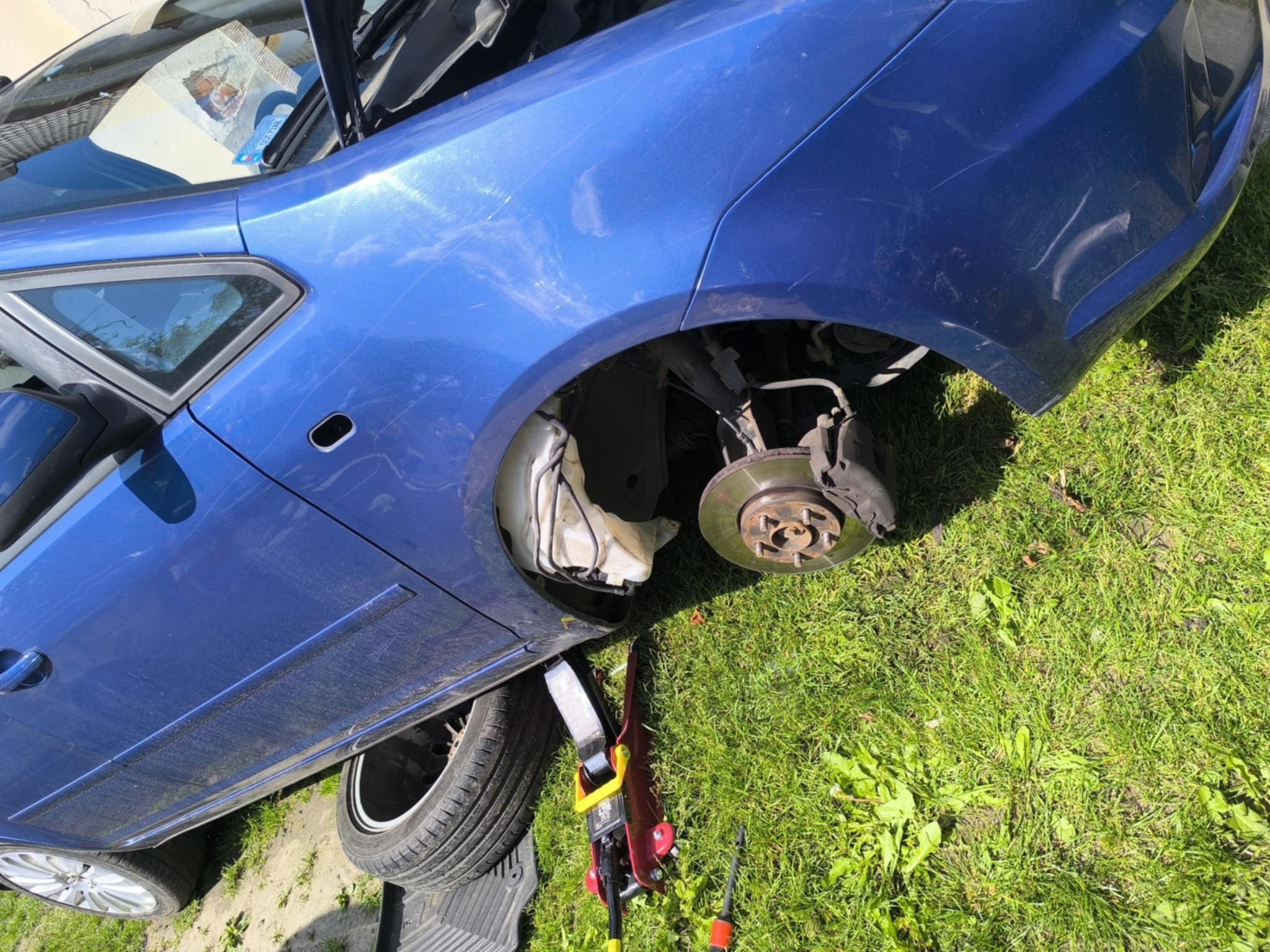 Niebieski samochód z podniesioną maską, bez koła, podnośnik na trawie. Widoczne tarcze hamulcowe i elementy zawieszenia. Prace naprawcze w toku, w plenerze.