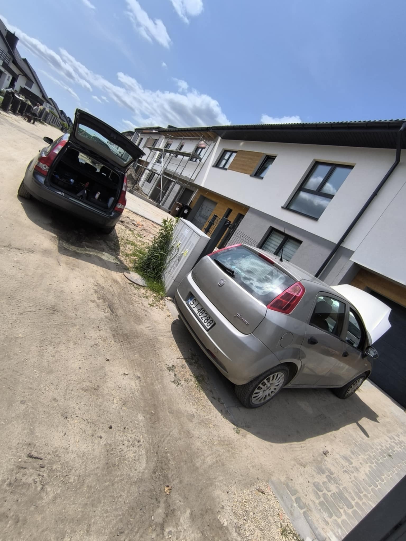 Dwa samochody: szary Fiat Punto z otwartą maską i czarny Volvo z otwartym bagażnikiem, stojące na żwirowej drodze przed nowymi domami.