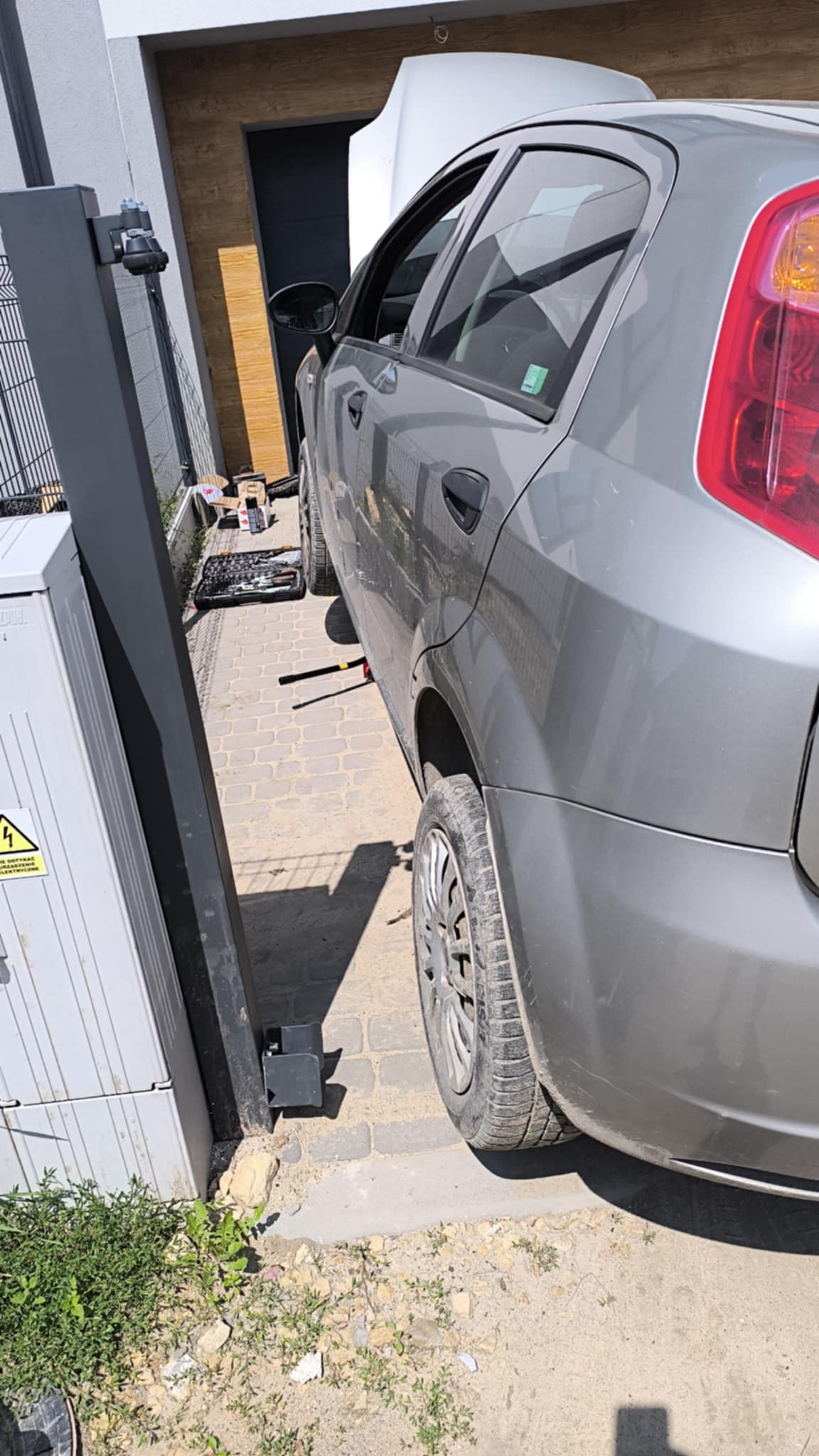 Szary samochód osobowy z otwartą maską, uniesiony na podnośniku, narzędzia w pobliżu. Auto zaparkowane na brukowanej nawierzchni, obok skrzynka elektryczna.