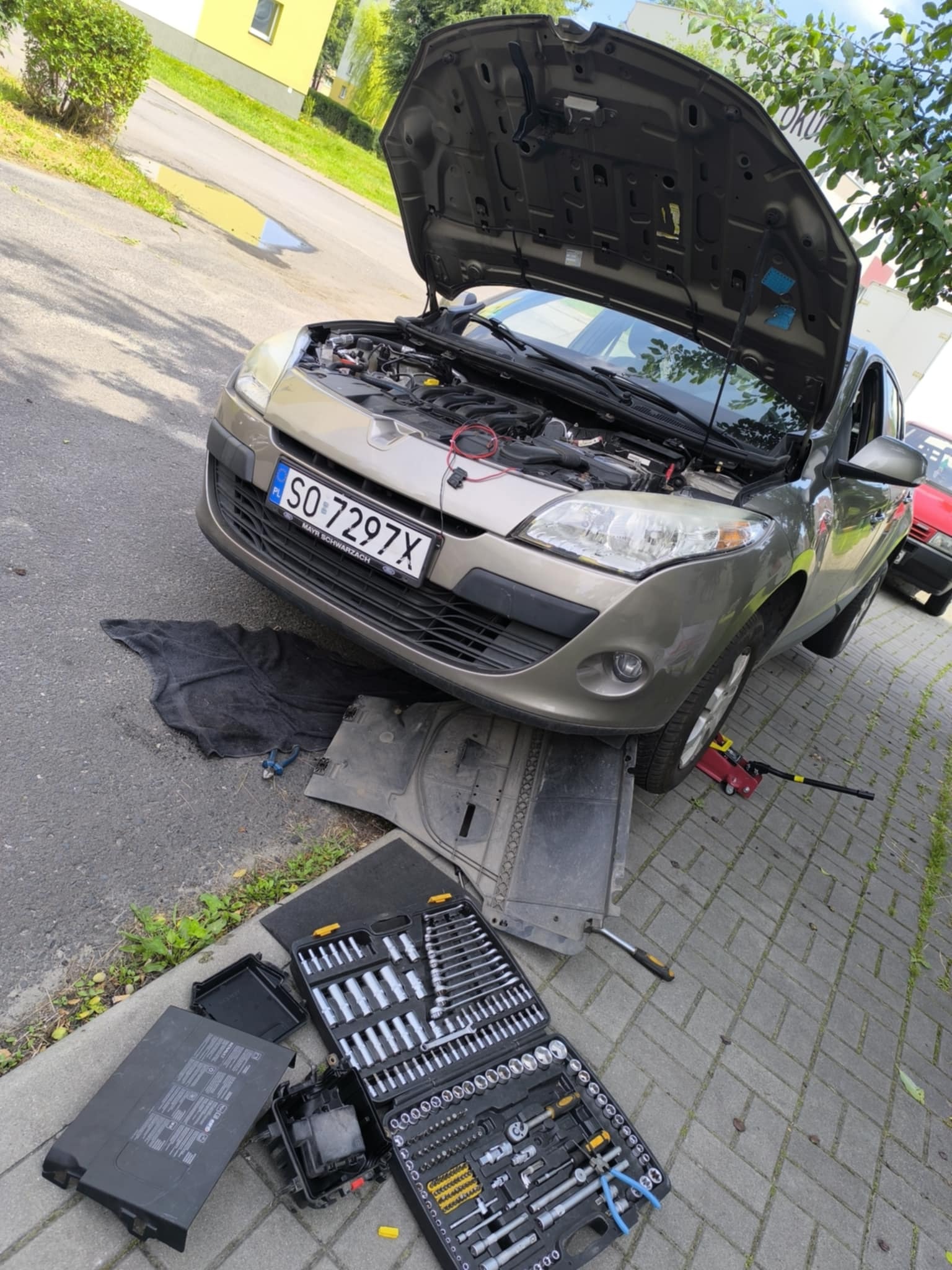 Beżowy samochód z otwartą maską na chodniku. Widoczny zestaw narzędzi, podnośnik i czarna mata. Tablica rejestracyjna SO 7297X. Prace naprawcze na zewnątrz.