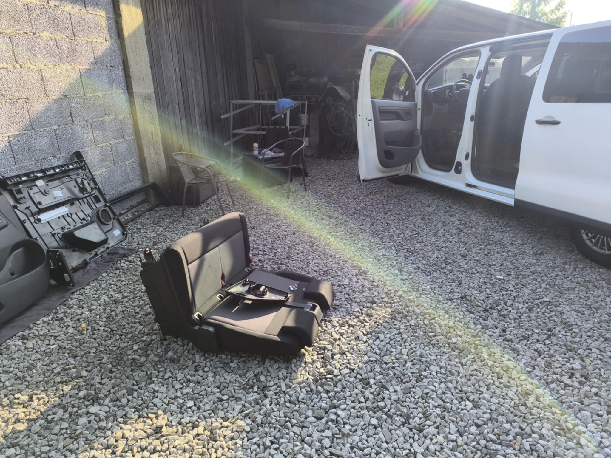 Wnętrze białego vana z otwartymi drzwiami; na zewnątrz, na żwirowym podłożu, leży wymontowany fotel i elementy tapicerki. Widoczne narzędzia w tle.