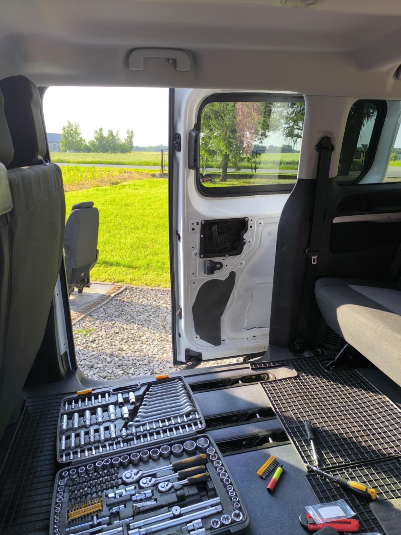 Wnętrze białego vana z otwartymi drzwiami, widoczny zestaw narzędzi i częściowo zdemontowane elementy. W tle zielone pole i budynki.