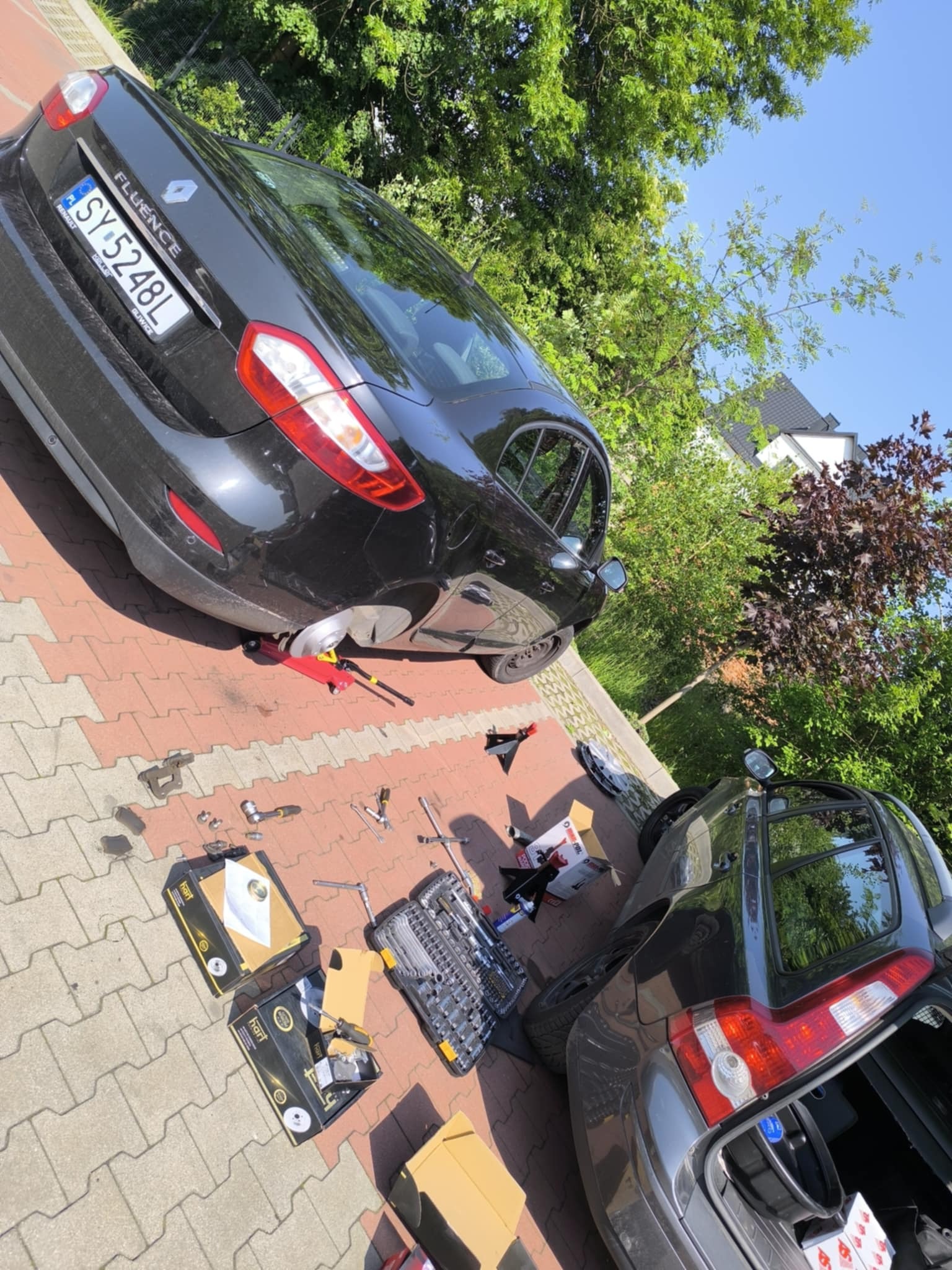 Czarny Renault Fluence na podnośniku, brak koła. Rozłożone narzędzia i części samochodowe obok auta. Dwa samochody na kostce brukowej, wśród zieleni.