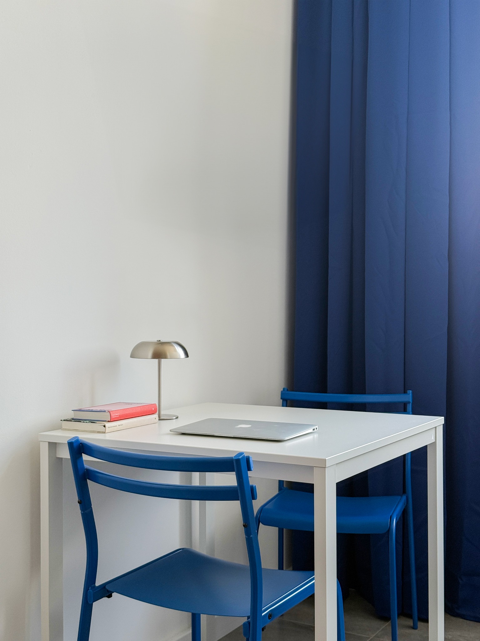 Białe biurko z laptopem, lampą i książkami, otoczone niebieskimi krzesłami i zasłoną. Minimalistyczna aranżacja wnętrza w chłodnej tonacji.