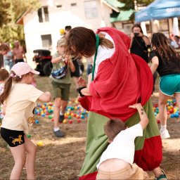 nie.movie - Animator w barwnym stroju otoczony dziećmi podczas zabawy z kolorowymi piłkami na trawie. W tle budynek i namiot.