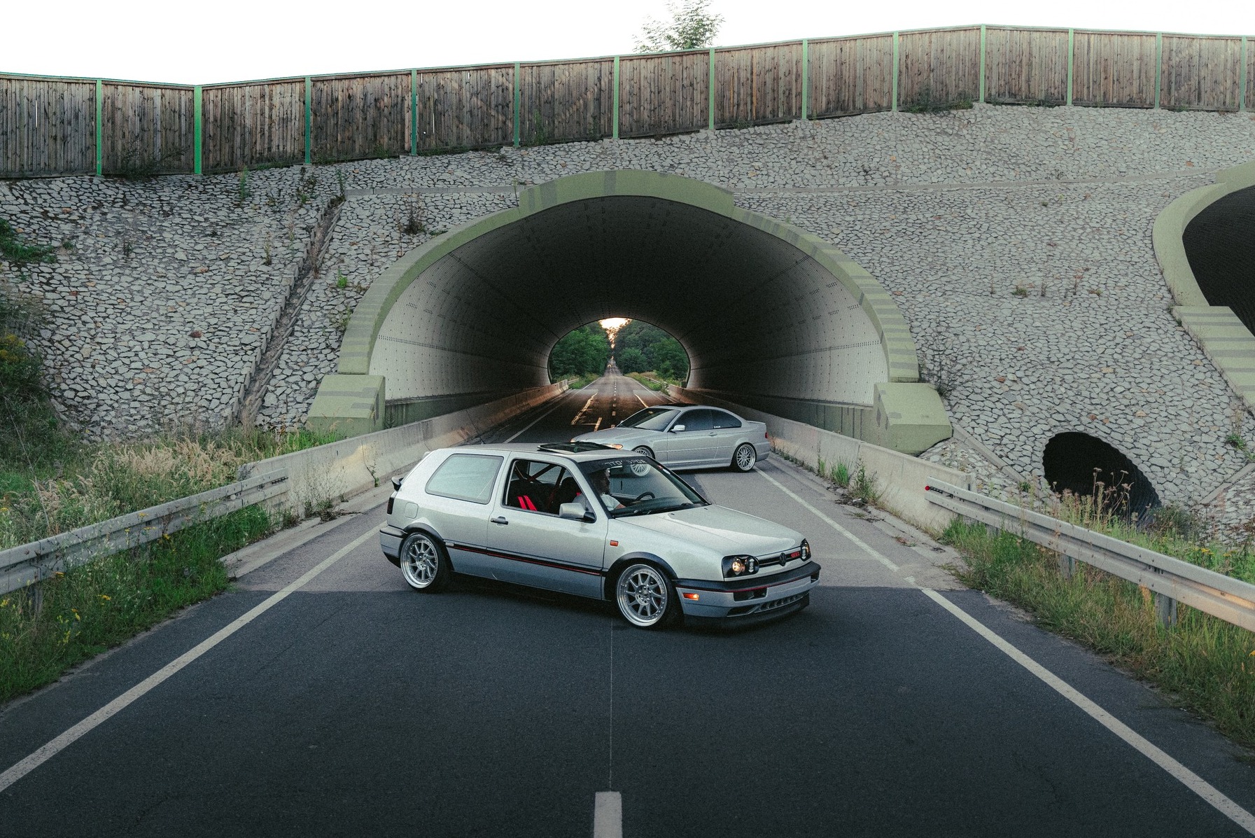 Dwa srebrne samochody, Golf III i BMW E46, stoją na asfalcie przed tunelem. Golf z otwartym szyberdachem, felgi aluminiowe. Ujęcie z niskiej perspektywy, lekko z dołu.