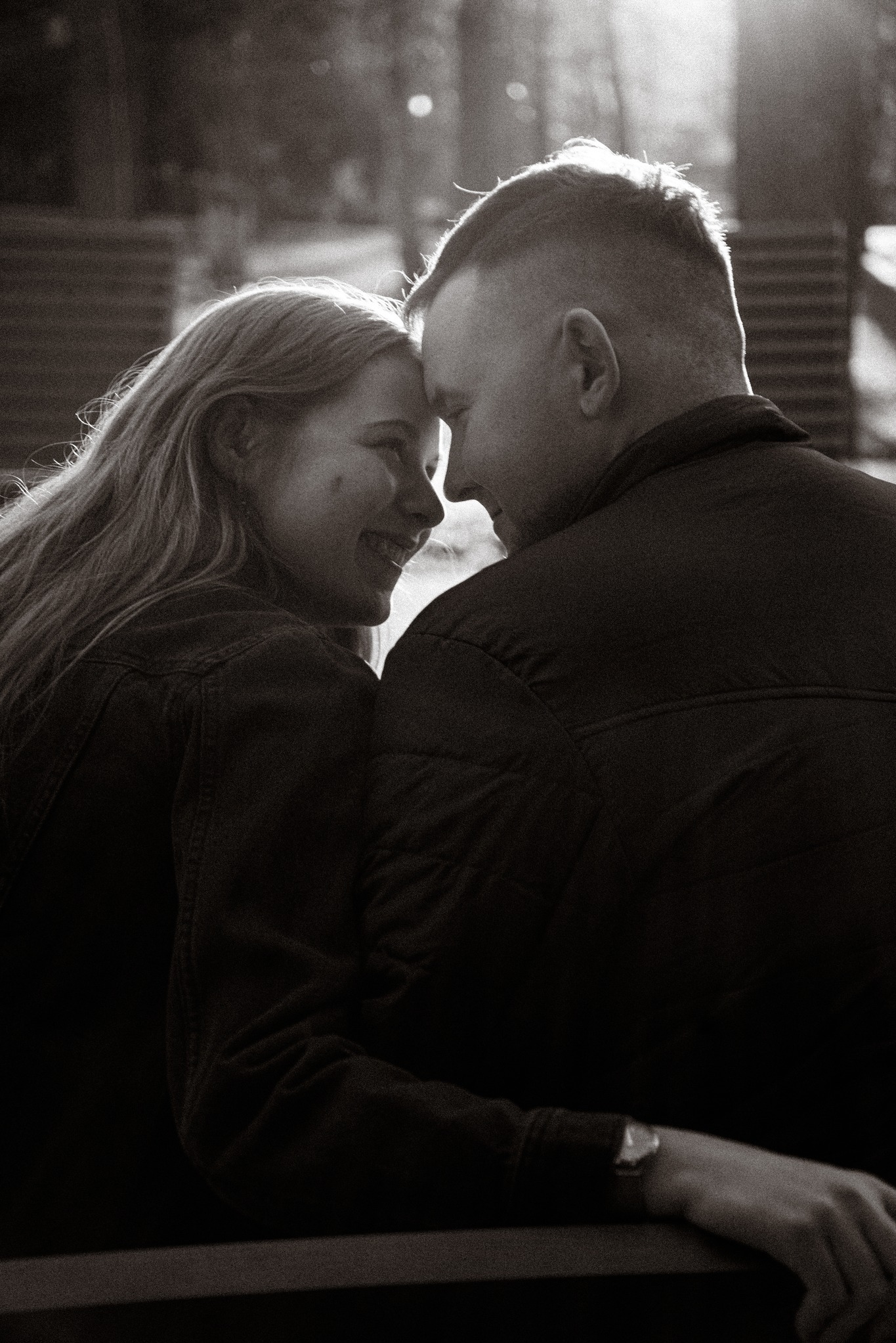 Czarno-białe zdjęcie narzeczeńskiej pary, stykającej się czołami i uśmiechającej do siebie, w plenerze. Widoczny zegarek na nadgarstku mężczyzny.