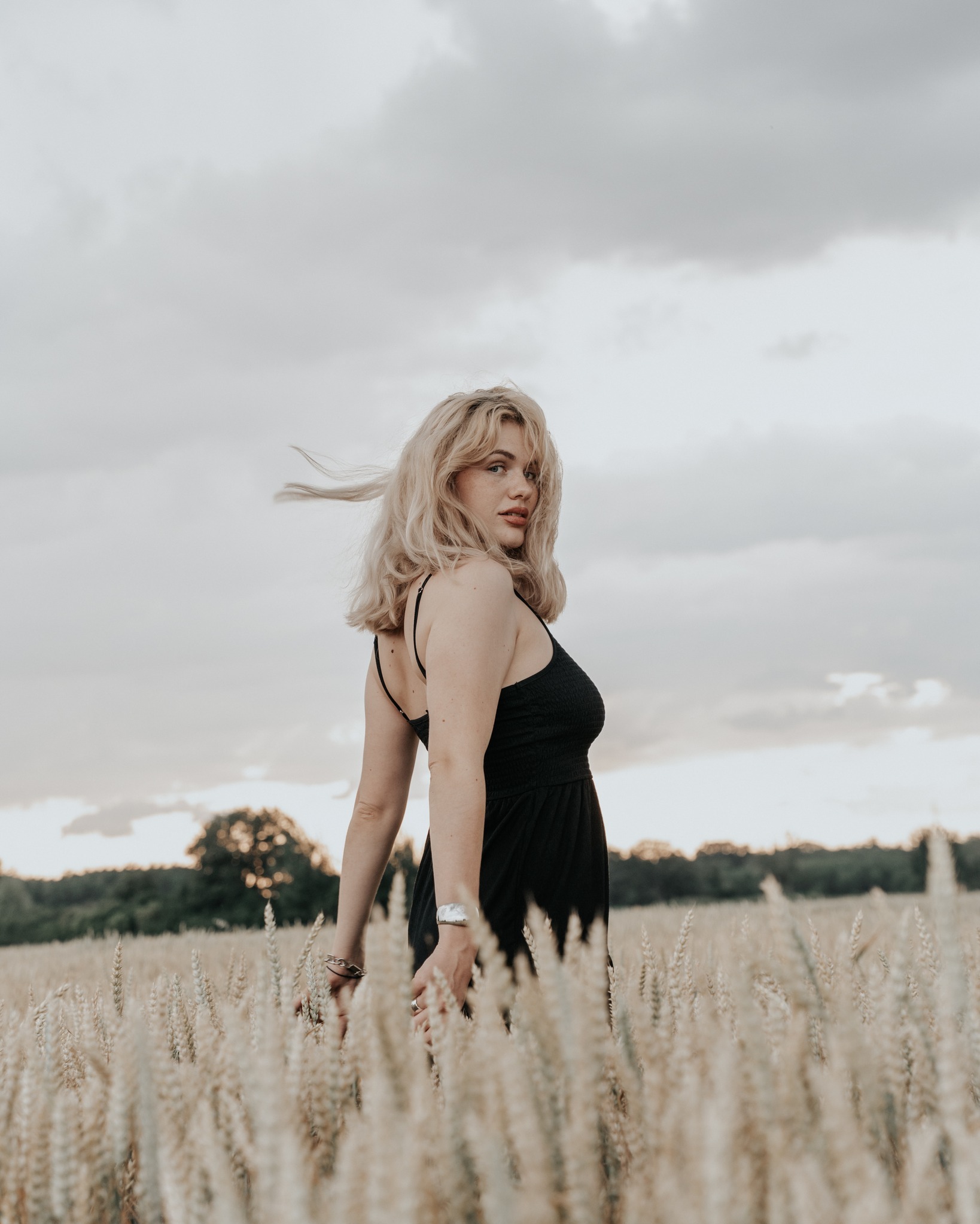Portret kobiety w czarnej sukience, odwróconej przez ramię na tle pola pszenicy i pochmurnego nieba, z wiatrem we włosach i delikatnym oświetleniem.