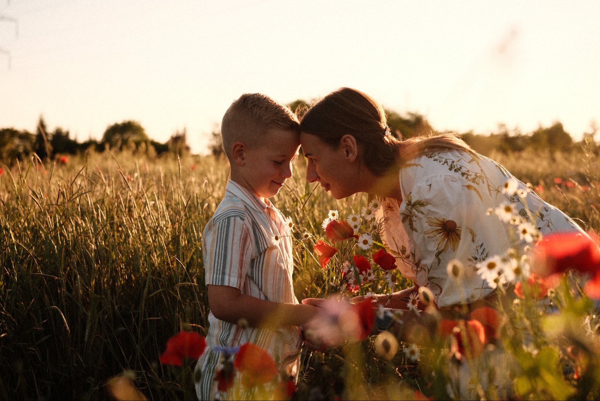 Matka i syn w letnim polu maków, stykają się czołami w czułym geście. Ciepłe światło zachodzącego słońca podkreśla bliskość i naturalność sceny.