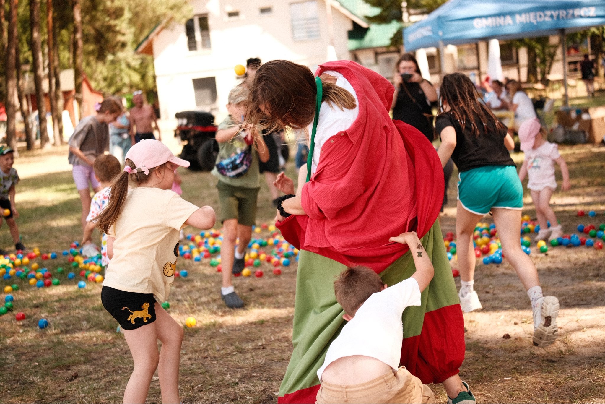 Animator w barwnym stroju otoczony dziećmi podczas zabawy z kolorowymi piłkami na trawie. W tle budynek i namiot.