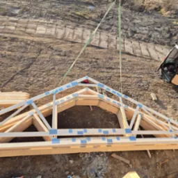 Gotowa więźba dachowa z drewnianych belek, zabezpieczona taśmą, podnoszona dźwigiem na placu budowy. W tle widoczny bus z otwartymi drzwiami.