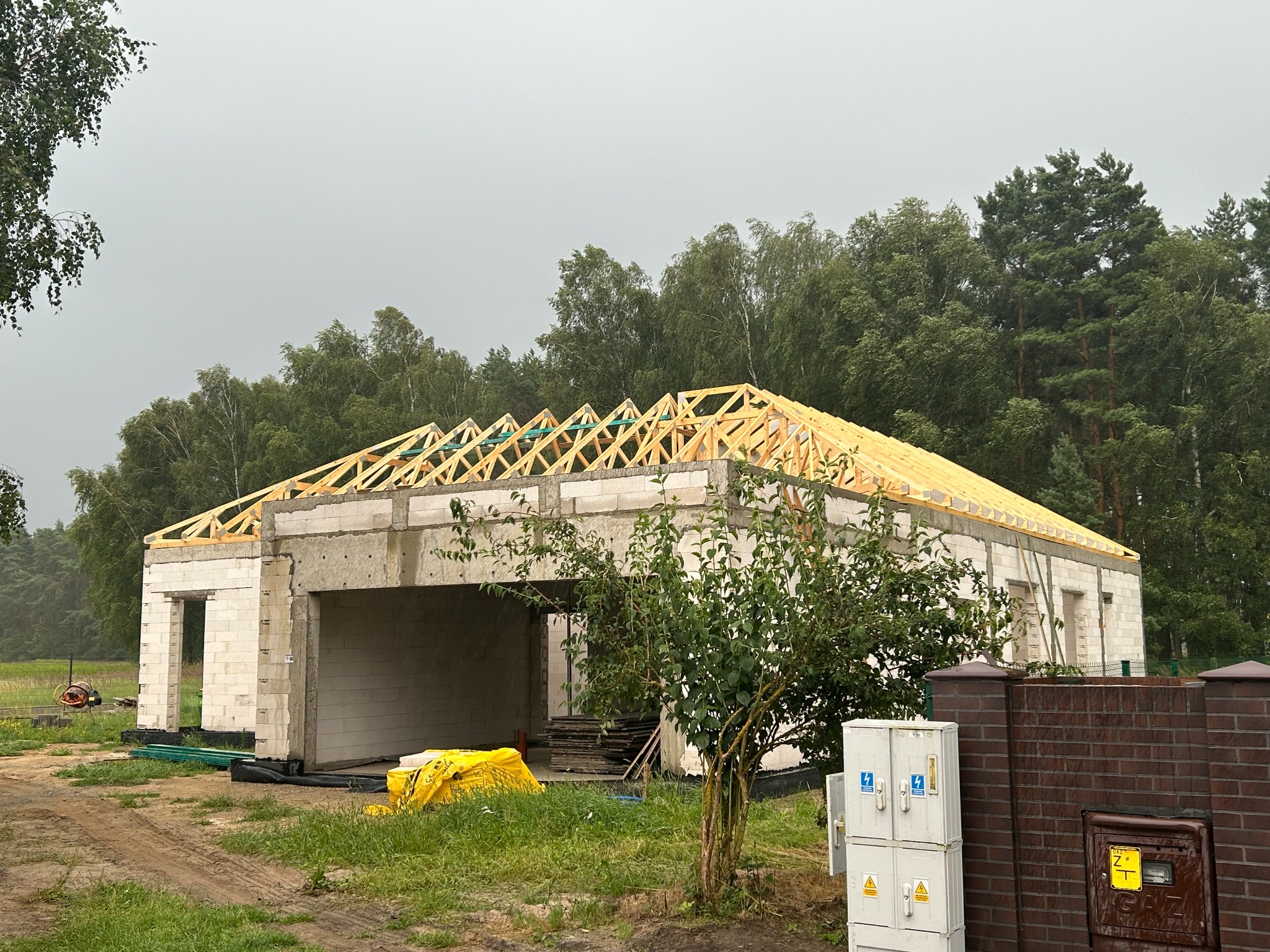 Budowa domu jednorodzinnego: surowy stan z częściowo wykonaną drewnianą więźbą dachową. Widoczne bloczki betonowe i elementy konstrukcyjne.