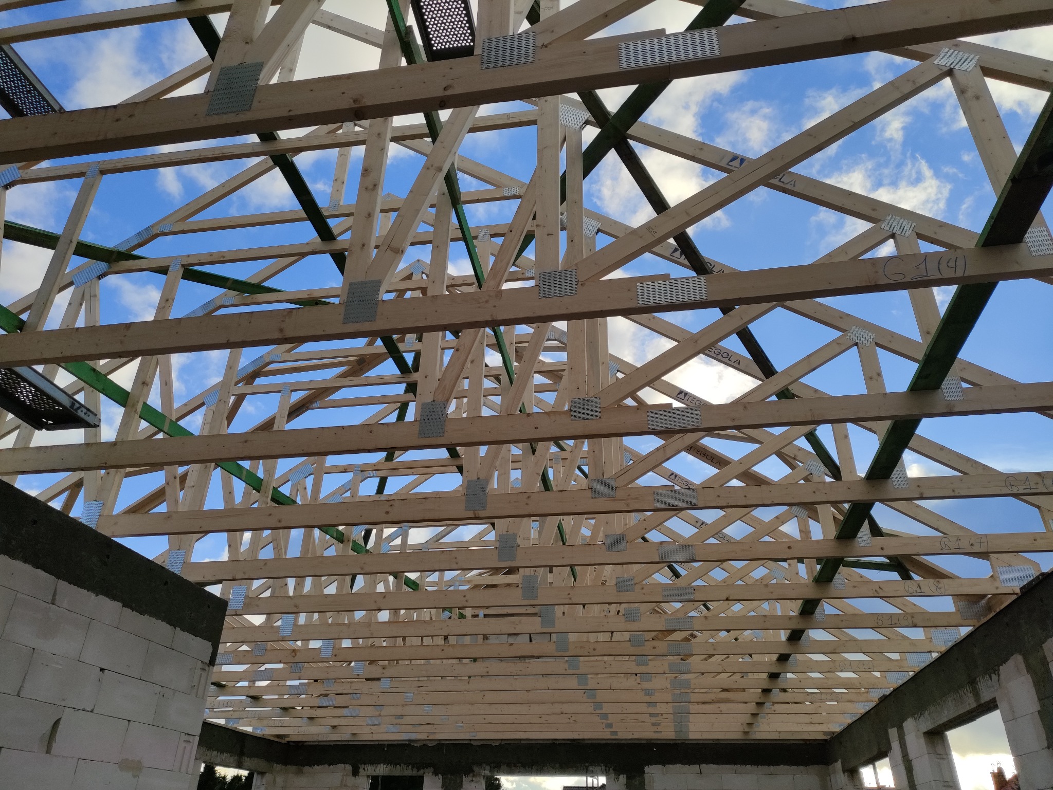 Drewniana konstrukcja więźby dachowej, widok z wnętrza budynku, z widocznymi elementami mocującymi i fragmentami ścian z bloczków. Błękitne niebo w tle.