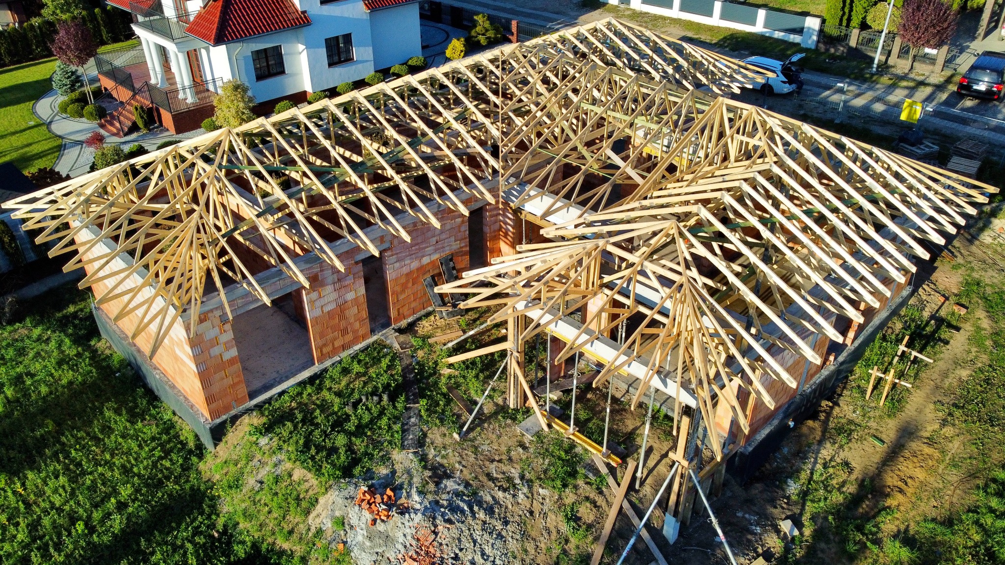 Wznoszenie konstrukcji drewnianej dachu na murowanym budynku z czerwonej cegły, widok z góry, zielona trawa wokół, częściowo zadaszony budynek w tle.