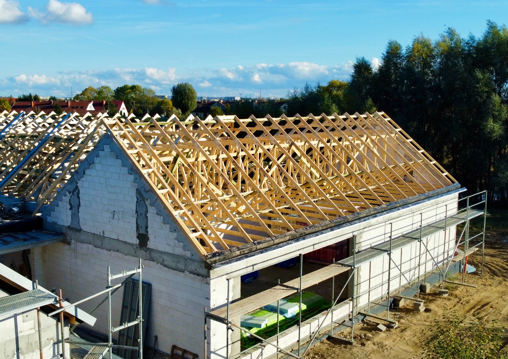 Wznoszenie drewnianej konstrukcji dachu na murowanym budynku w trakcie budowy. Widoczne rusztowania i składowane materiały budowlane. Słoneczny dzień.