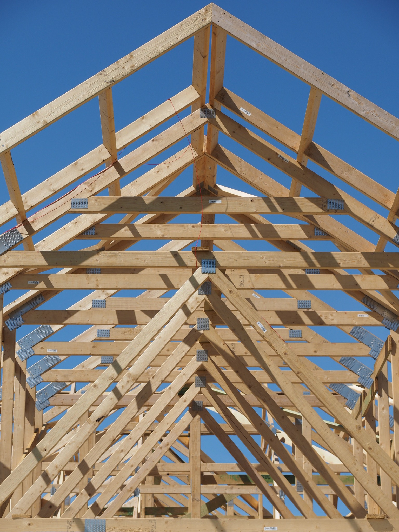 Drewniana konstrukcja dachu z widocznymi łącznikami metalowymi, na tle błękitnego nieba. Układ symetryczny, w centralnej części kadru. Jasne drewno, wyraźne detale.