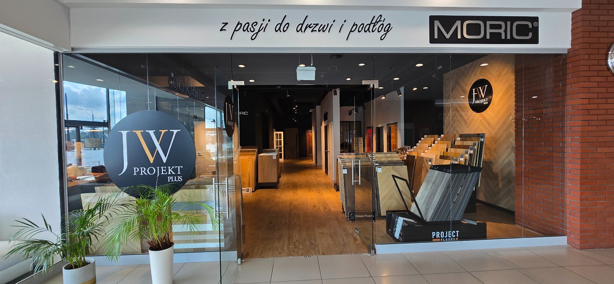 Salon z ekspozycją drzwi i podłóg drewnianych, widoczne próbki paneli ułożone na stojakach, logo firmy na szybie i nad wejściem. W głębi widoczne drzwi.