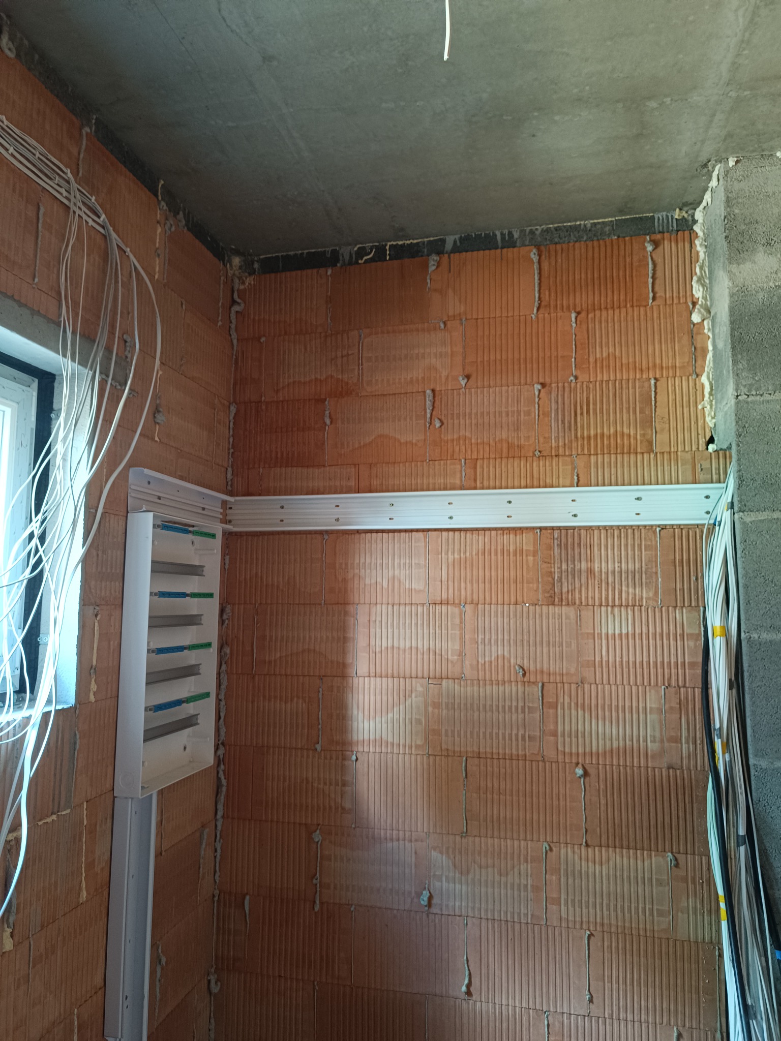 Nierówna ściana z cegły z zamontowaną białą skrzynką elektryczną i wiązkami kabli. Widoczne przewody instalacji elektrycznej przed tynkowaniem.