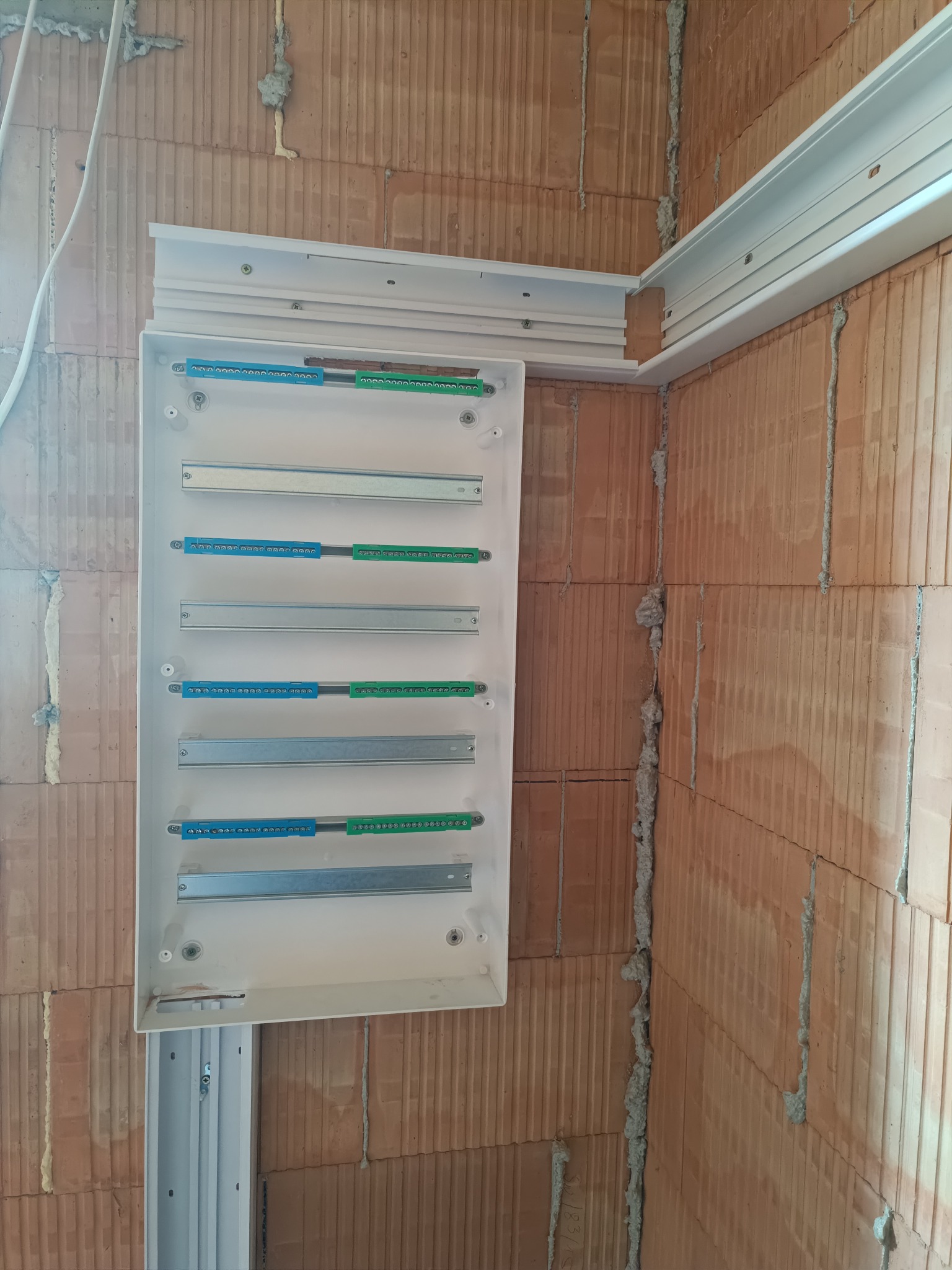 Otwarta skrzynka elektryczna z listwami zaciskowymi na ceglanej ścianie. Widoczne przewody i elementy montażowe. Etap prac instalacyjnych.