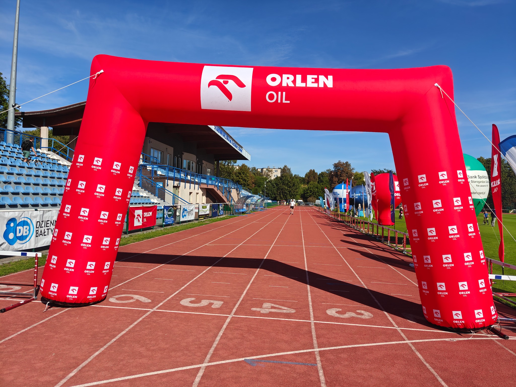 Czerwony, dmuchany łuk startowy Orlen Oil na bieżni stadionu lekkoatletycznego w słoneczny dzień. Widoczne trybuny i flaga. Perspektywa z linii startu.
