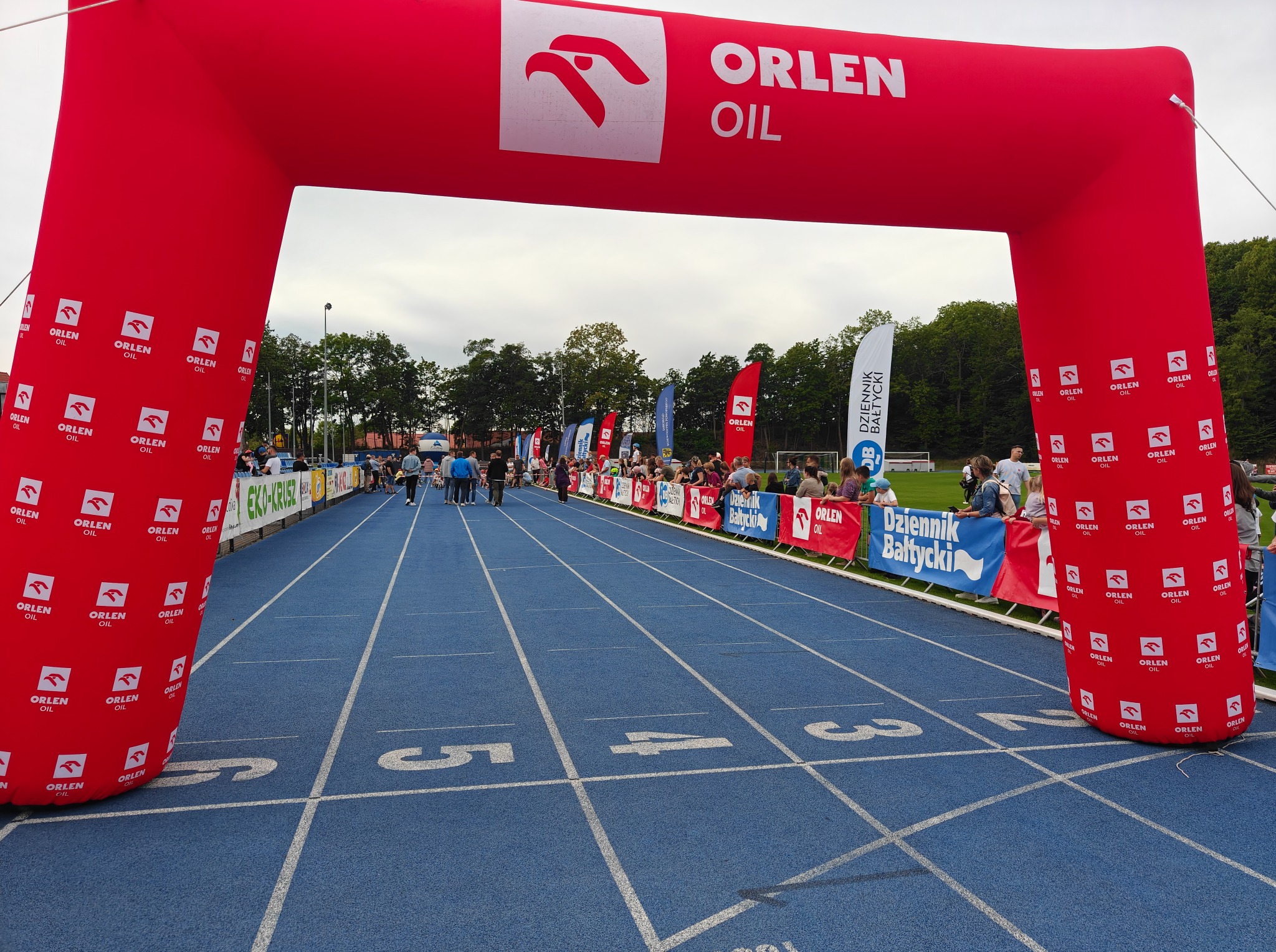 Brama startowa Orlen Oil na stadionie lekkoatletycznym z niebieską bieżnią i numerami torów. W tle widoczni ludzie i flagi sponsorskie.