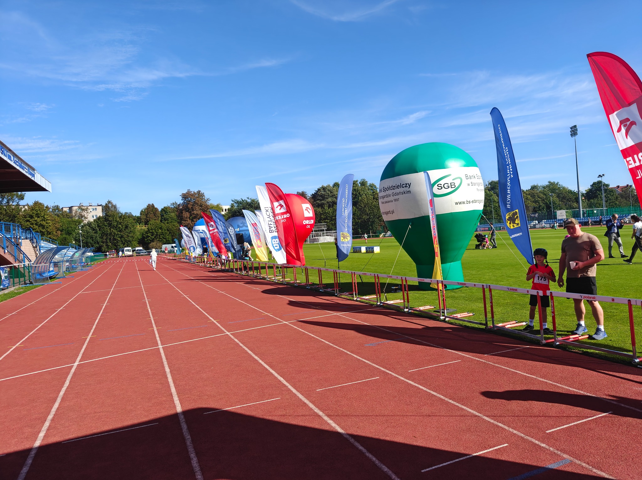 Bieg na stadionie: czerwona bieżnia, kolorowe flagi reklamowe, dmuchany balon SGB. Widoczni uczestnicy i kibice. Słoneczny dzień, błękitne niebo.