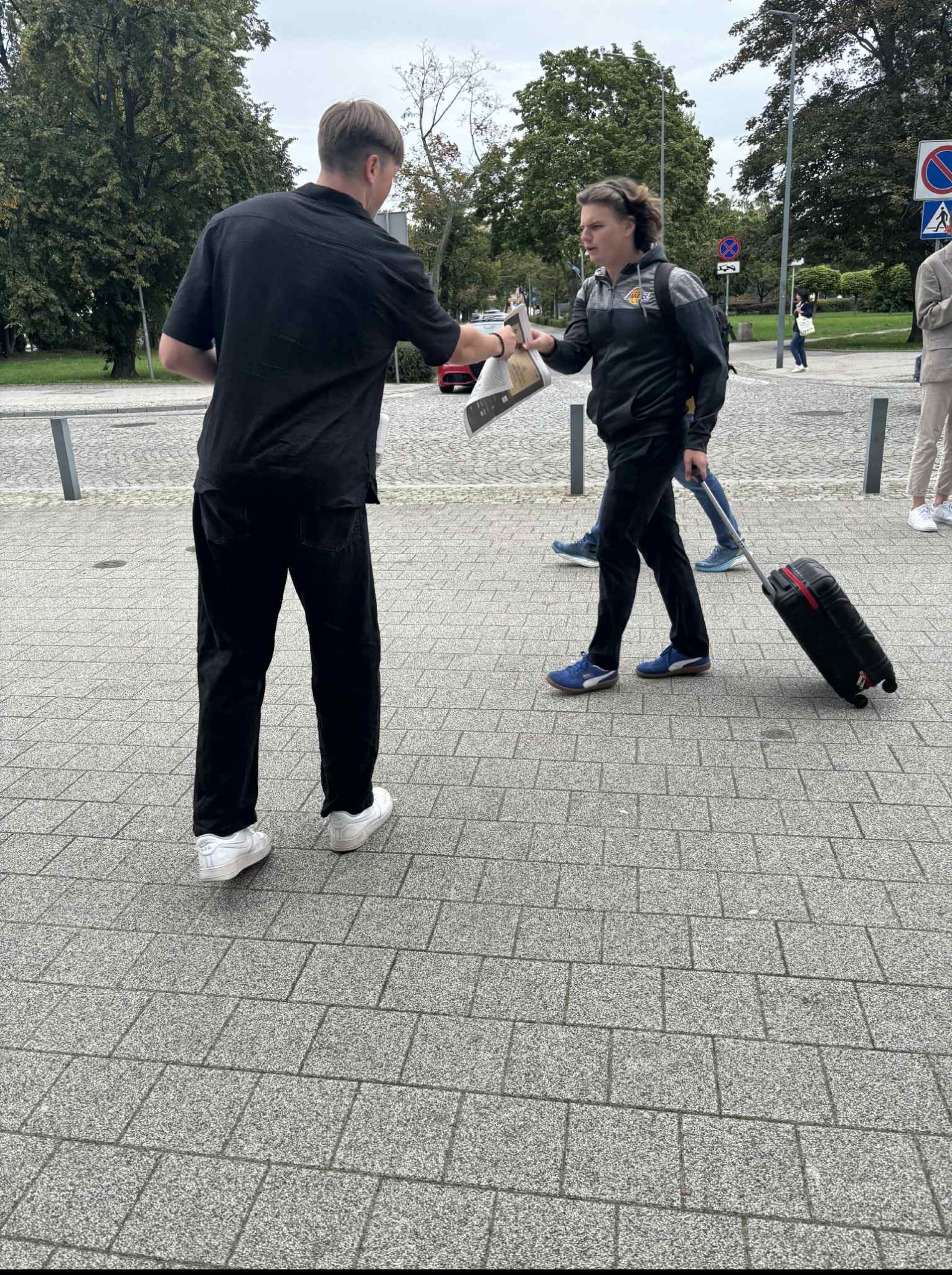 Mężczyzna w czarnej koszuli wręcza ulotki kobiecie z walizką na brukowanej ulicy, prawdopodobnie w pobliżu miejsca wydarzenia.