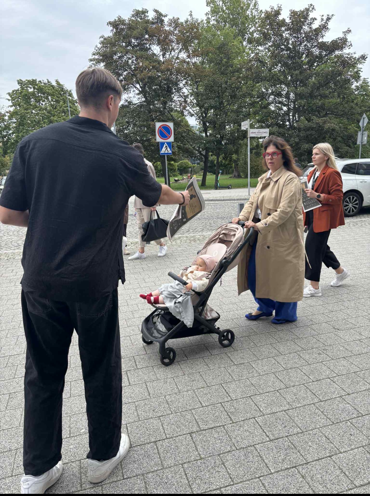 Mężczyzna wręcza ulotkę kobiecie z wózkiem dziecięcym na tle ulicy z zielenią miejską. Reklama zewnętrzna, dystrybucja materiałów promocyjnych.